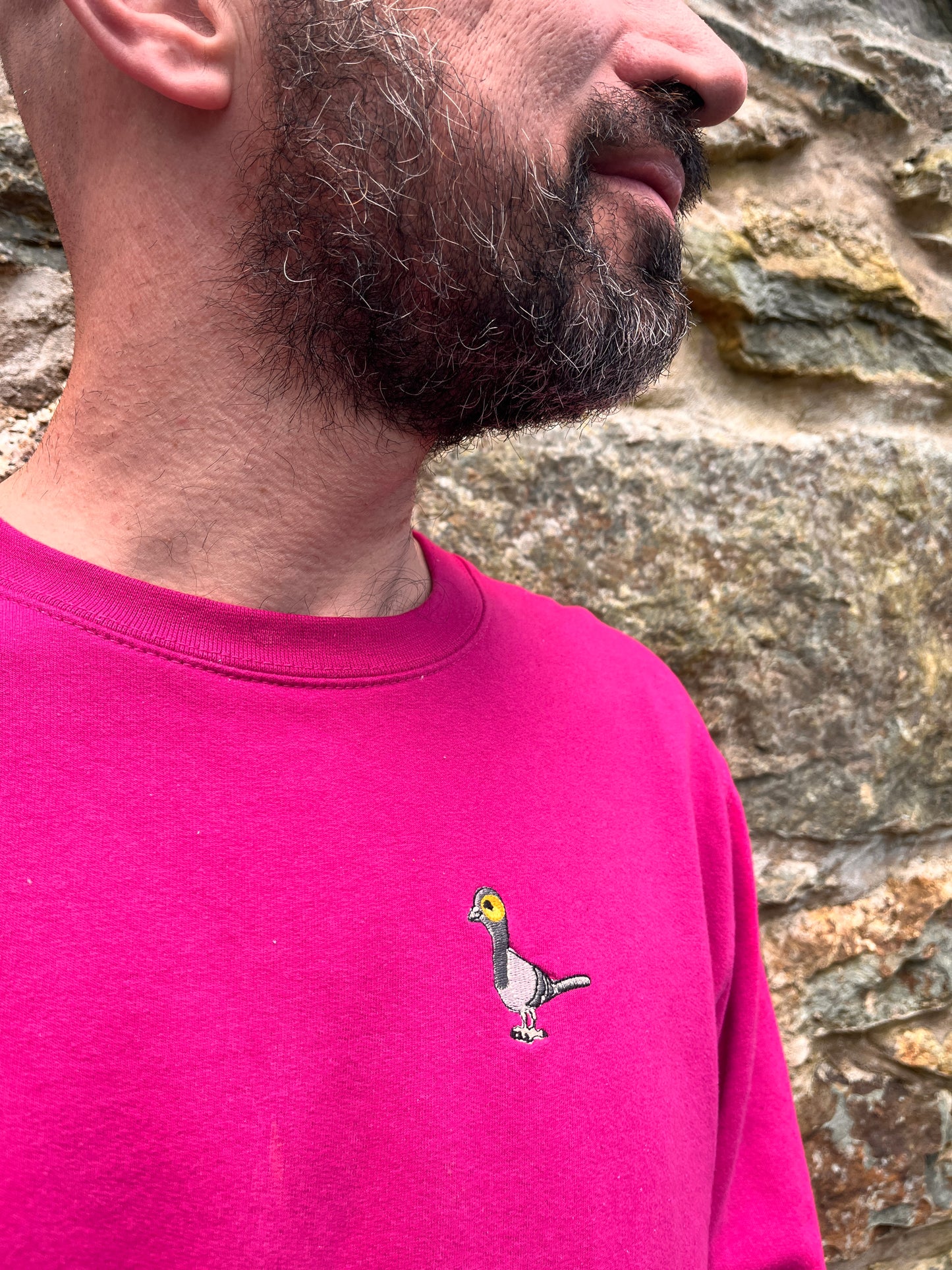 Person wearing a pink shirt with a small embroidered pigeon logo against a rocky background